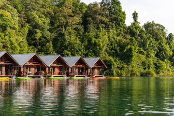 Fototapeta premium Row of wooden overwater bungalows on a tranquil lake, with lush green jungle mountains in the background under soft daylight.