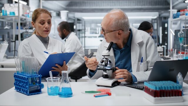 Laboratory technician examining biopsy slide while colleague documents diagnostic findings on clipboard. Elderly man identifies disease markers in clinical lab with coworker taking notes, camera B