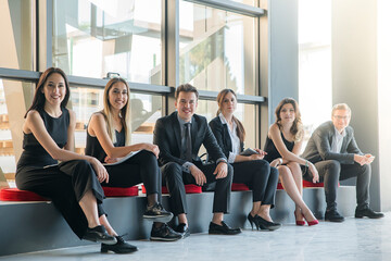 Fototapeta premium a group of smartly dressed, young professionals are sitting on a bench with red cushions in a modern office lobby, smiling warmly at the camera.