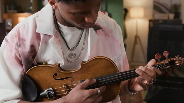 Tilt up shot of young adult African American male recording artist strumming strings of violin while experimenting with performance techniques in home studio