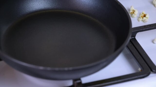 Close-up of an empty non-stick pan on a gas stove. Empty black frying pan with a non-stick coating resting on a modern white gas cooktop in a kitchen, with some popcorn kernels