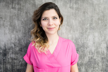 Fototapeta premium Smiling young woman in pink scrubs standing against gray background