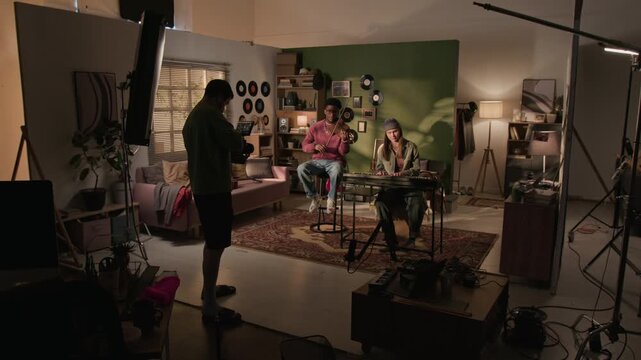 Wide backstage shot of camera operator shooting live session of two young adult musicians performing violin and synth in home studio setting