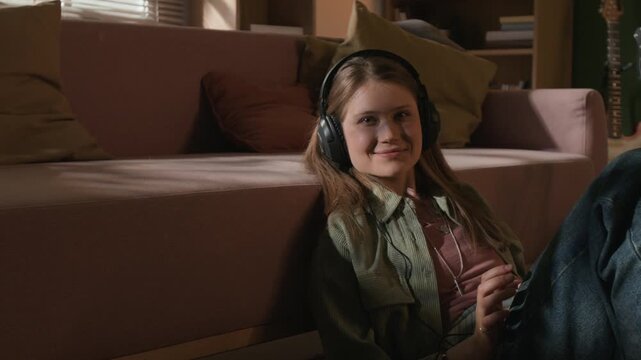 Medium portrait shot of smiling young adult female music producer wearing headphones listening to recorded demo while sitting in home studio