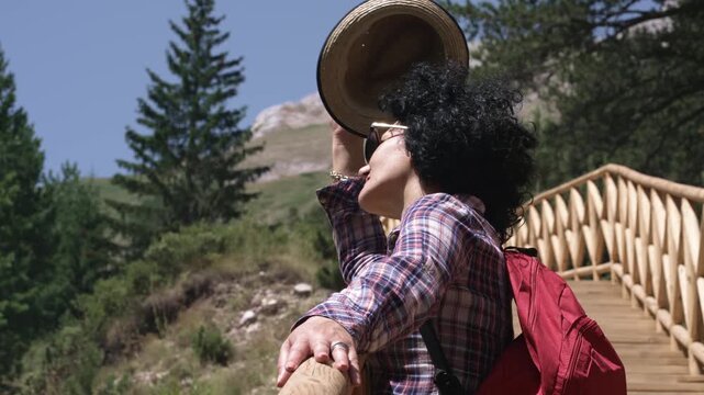 Hiker woman wearing a hat and a red backpack is standing on a wooden bridge. She is  looking out over a beautiful mountain range