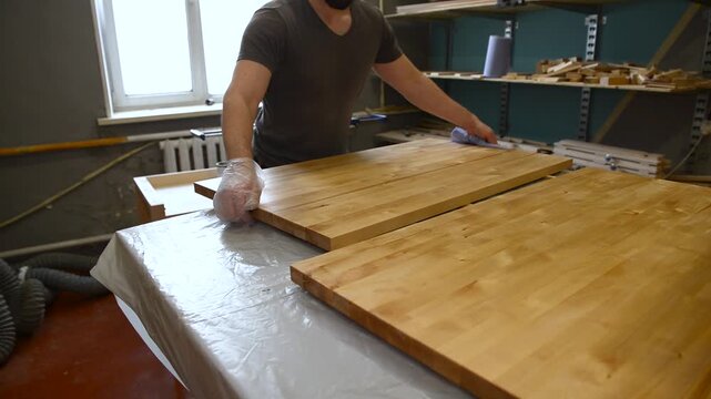 Professional carpenter in protective gloves working in workshop with wooden products, joiner applying gold colour paint on surface of raw wooed plank, worker varnishing wood for further furniture
