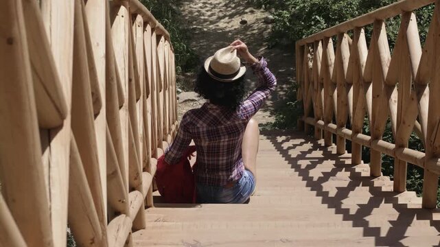 Hiker woman wearing a hat sits on a wooden staircase. She is holding a red backpack. The scene is peaceful and serene, with the woman enjoying a moment of relaxation