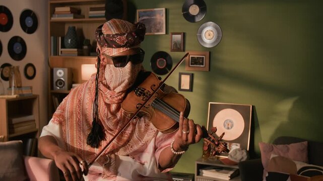 Medium shot of young adult Black male musician wearing Muslim traditional head covering playing violin in living room decorated with vinyl records and gold award plaque