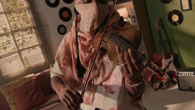 Push in shot of Black male musician wearing desert scarf covering face playing violin in duo with masked woman performing synth during band rehearsal in living room, Dutch tilt