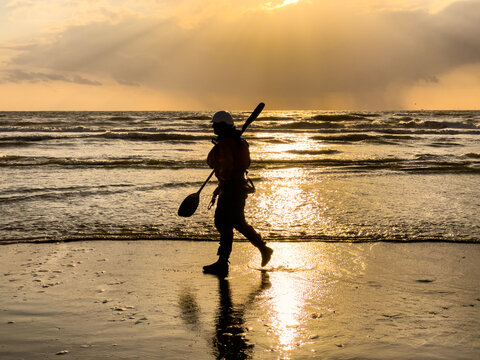 View of a person in silhouette carrying a paddle along the shoreline of Strand Wijk aan Zee during a golden sunset Wijk aan Zee, North Holland, Netherlands.