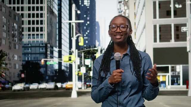 Black smiling interviewer filming news segment in city center using mic. Happy media professional on city sidewalk using microphone to produce reportage for local television station.