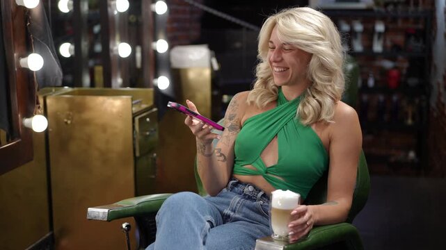 Joyful blonde woman with tattoos smiling while scrolling on her phone. She is sitting in a make-up room, holding a latte coffee