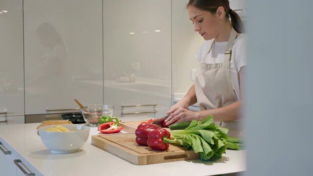 Woman in apron chopping red bell pepper on wooden cutting board. Home cook preparing vegetables for healthy meal in modern white kitchen. Culinary process of slicing fresh organic produce for dinner