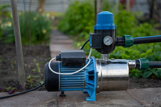 Surface water pump with automatic pressure control switch and gauge installed on a garden path tile, connected to black irrigation pipes with green fittings.