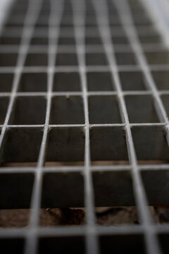 Close up perspective view of a galvanized steel drainage grate with selective focus, showing a repetitive geometric metal grid pattern fading into blurred background.