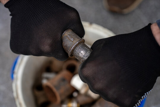 Hands in black protective gloves trying to unscrew or assemble an old rusty metal threaded pipe joint over a bucket containing plumbing scrap.