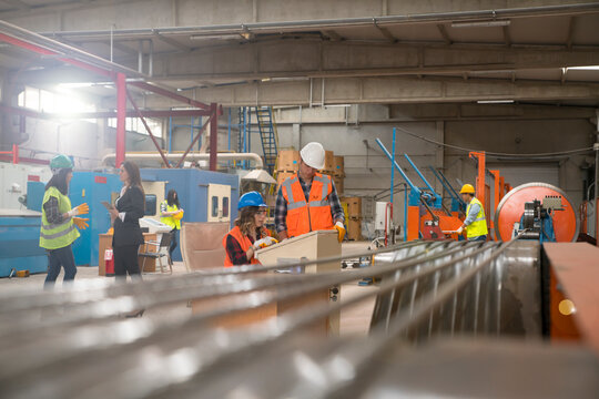 Workers in hard hats and high-visibility vests are engaged in various activities on the factory floor, including inspecting materials and collaborating with colleagues.