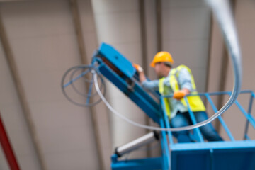 Fototapeta premium A construction worker wearing a yellow hard hat and high-visibility vest operates an aerial lift, signaling with his hand. The scene suggests a workplace safety or industrial setting.