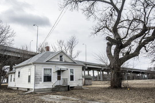 View of a small white house and a large bare tree with a concrete highway overpass in the background under an overcast sky in Lincoln, Nebraska, United States.