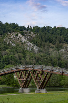 View of the arched wooden bridge over the river with lush green forests and rocky cliffs under a soft sky in Essing, Bayern, Germany.