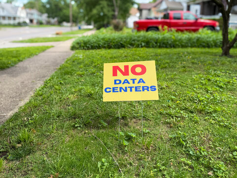 Close-up shot of a yellow and red "No Data Centers" grassroots protest sign placed in a green grass lawn. Selective focus with a blurred city neighborhood background.