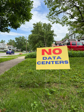 Vertical view of a yellow and red "No Data Centers" political protest sign positioned in the front yard grass of a city neighborhood. Clean composition with copy space.