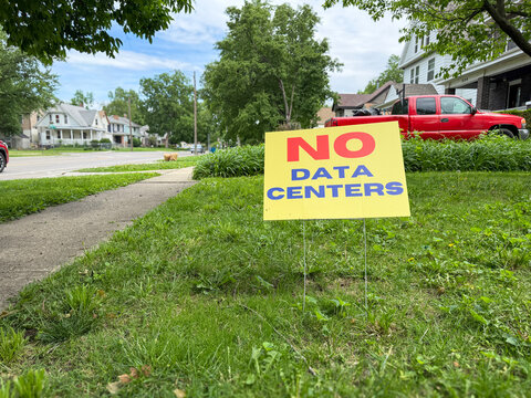 Residential Front Yard with No Data Centers Sign. A yellow and red "No Data Centers" political protest sign positioned in the front yard of a city neighborhood. Clean composition with copy space.