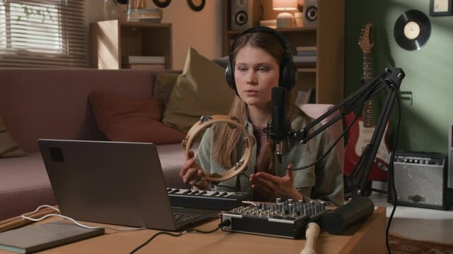 Medium shot of young adult female musician recording tambourine jingles, processing sounds into samples while creating rhythmic patterns in DAW on laptop in home studio