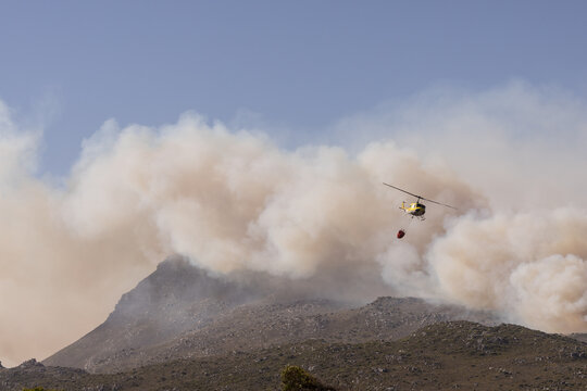 View of a yellow firefighting helicopter carrying a water bucket near thick smoke rising from a wildfire on the slopes of Table Mountain Cape Town, Western Cape, South Africa.