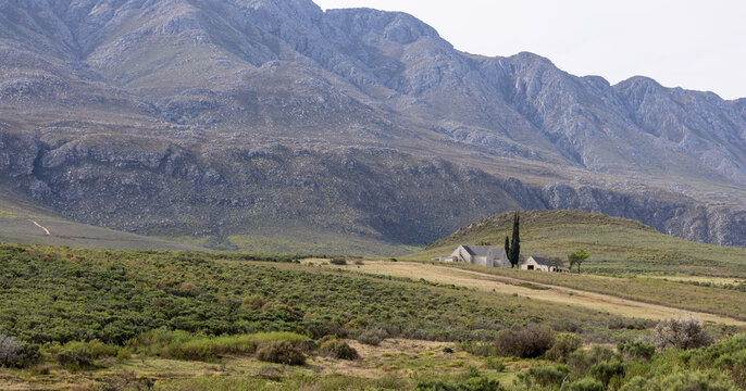 View of the rugged Swartberg Mountains towering over a small farmhouse nestled among green shrubs and arid landscape in Swartberg, Western Cape, South Africa.