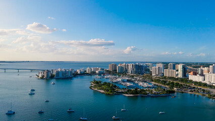 Fototapeta premium High-angle aerial panorama of a luxury coastal city marina filled with white yachts and sailboats near high-rise skyline buildings and a long bridge