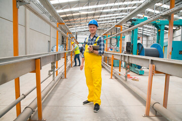 Fototapeta premium A smiling industrial worker in a blue hard hat and yellow overalls holds a clipboard on a factory floor with coworkers in the background, managing tasks and inspecting equipment.