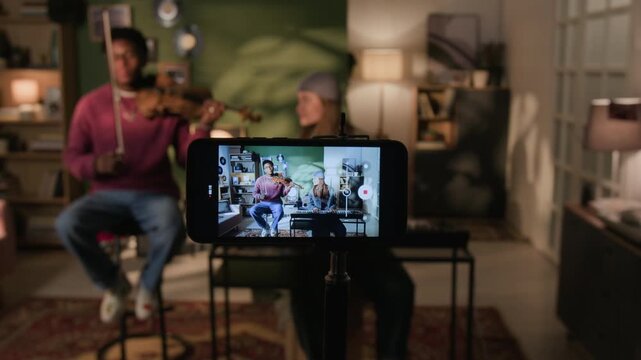 Close up shot of smartphone recording performance of two young adult musicians playing violin and synth in living room creating social media content