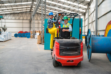 Two men wearing hard hats ride a forklift in a warehouse, one sitting and one standing. They navigate through a storage area with large pipes and industrial equipment. © bulentbaris