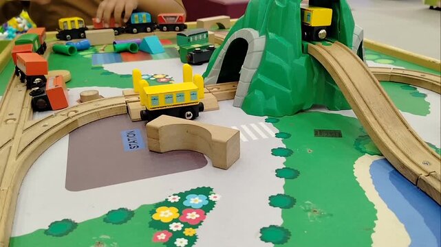 A boy's hand is playing with a children's toy train with carriages on wooden tracks in a playground.
