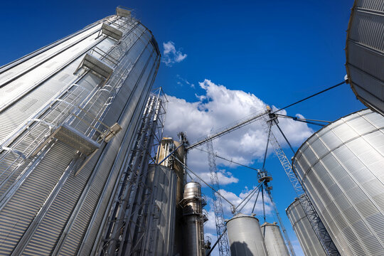 View of tall silver industrial grain silos and metal storage elevators under a bright blue sky with white clouds in Toronto, Ontario, Canada.