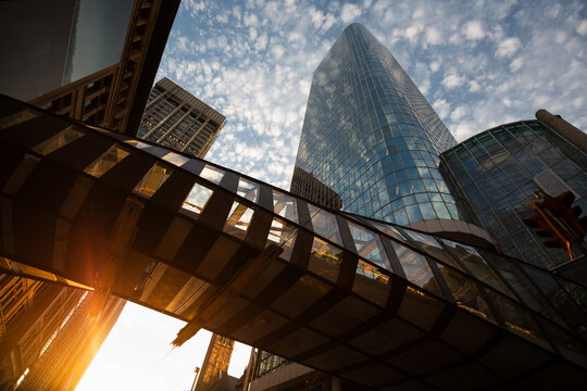 View of a modern glass skyscraper and a pedestrian skybridge under a blue sky with scattered clouds and bright sunlight flare in Toronto, Ontario, Canada.