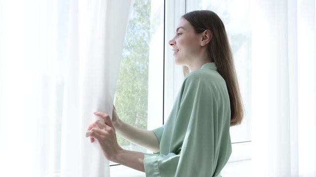 Smiling woman in bathrobe looking at window with beautiful curtains at home. Camera moving forward and backwards