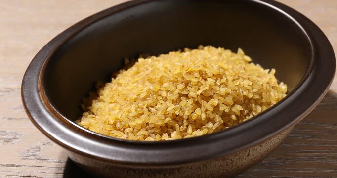 Pouring raw bulgur with spoon into bowl at wooden table, closeup
