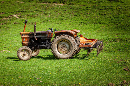 View of an orange tractor with a plow attachment parked on a lush green grassy hillside in Arang Kel, Azad Kashmir, Pakistan.