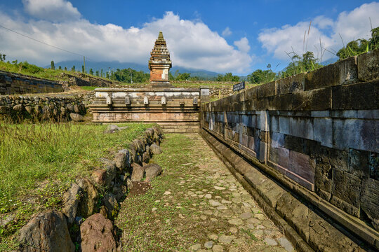 View of the ancient Situs Liyangan archaeological site featuring stone temple structures and terraces under a blue sky in Temanggung, Central Java, Indonesia.