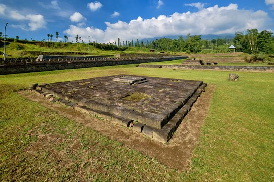 View of the ancient stone foundations of Situs Liyangan surrounded by green grass and stone walls under a blue sky in Temanggung, Central Java, Indonesia.