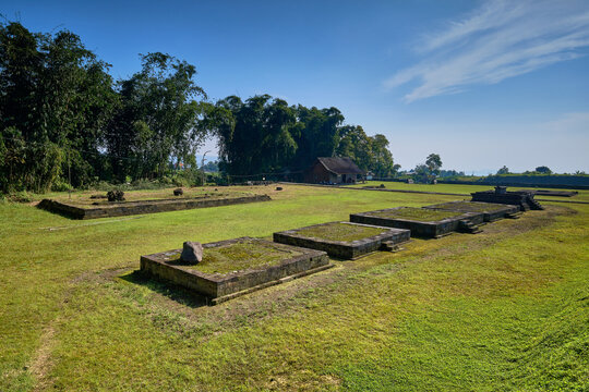 View of the ancient stone foundations of Situs Liyangan archaeological site surrounded by green grass and bamboo trees under a clear blue sky in Temanggung, Central Java, Indonesia.