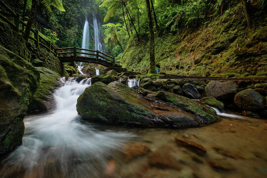 View of Jumog Waterfall cascading through lush tropical greenery with a wooden bridge over a rocky stream Karanganyar, Central Java, Indonesia.