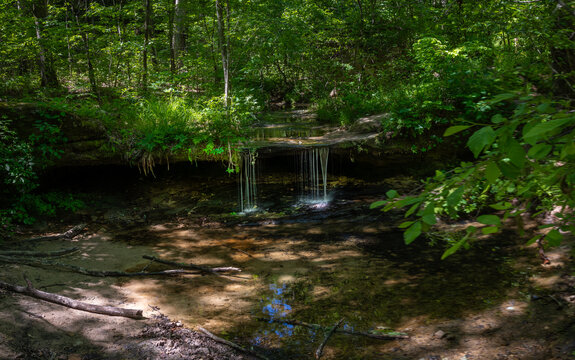 View of Pickle Springs Natural Area with a small waterfall cascading over a rock ledge into a clear pool surrounded by lush green forest New Offenburg, Missouri, United States.