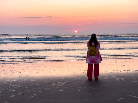 Wijk aan Zee, Netherlands - 06 June 2024: View of a person with a yellow backpack standing on Strand Wijk aan Zee watching the sunset over the North Sea with pink and orange sky.