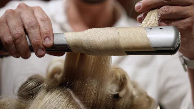 Close-up of a stylist using a flat iron to create elegant curls in a female client's long blonde hair. Professional haircare concept
