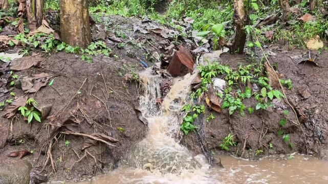 Dirty brown rainwater runoff flowing from the ground into a muddy river stream.