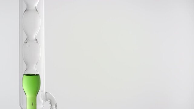 Pale green transparent fluid filling up a bulb lab condenser on the left side of the frame, right side for copy space on grey | Centella asiatica infusion cosmetic formulation design concept