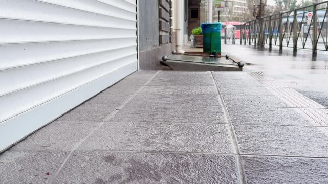 A white rolling shutter is pulled tightly shut at a shop entrance on a wet, gray day. The damp stone tiles and empty sidewalk create a quiet, lonely, and closed-off urban atmosphere.